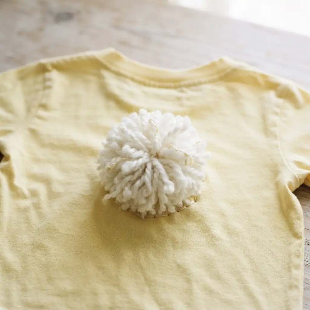 A pale yellow shirt with a large, fluffy white yarn pom pom sewn on the back.