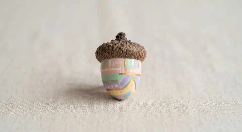 A shallow wooden bowl overflows with whole acorns painted in soft pastel shades of pink, green, and off-white.