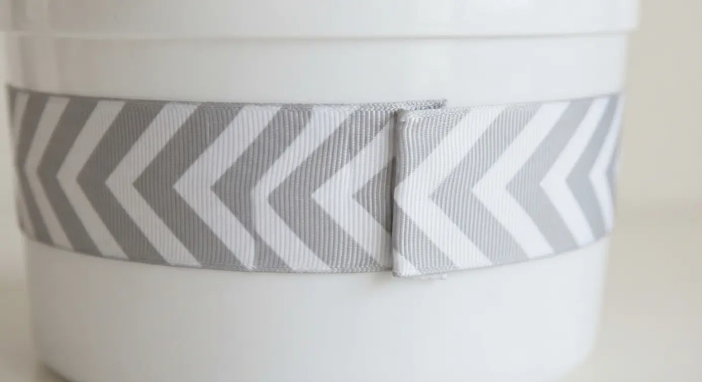 A clean white plastic pail is wrapped with a wide ribbon featuring a sharp gray and white chevron pattern.