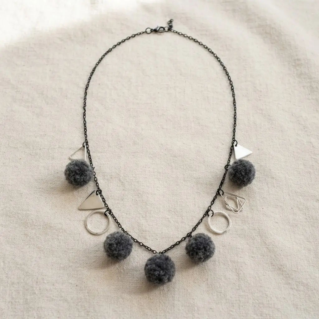 A contemporary necklace featuring silver geometric triangle and circle charms mixed with charcoal grey wool pom poms on a black chain.