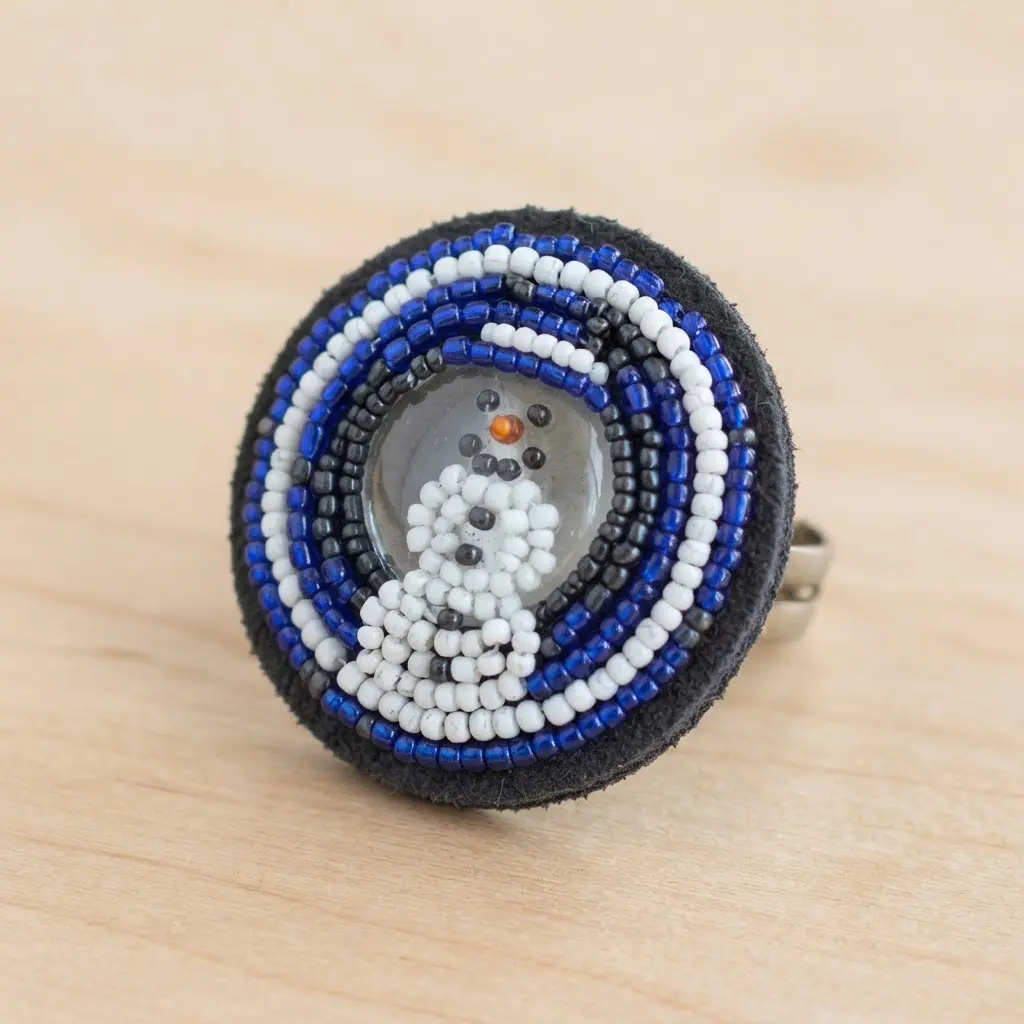 A flat ring features a snowman scene created with seed bead embroidery surrounding a central glass cabochon.