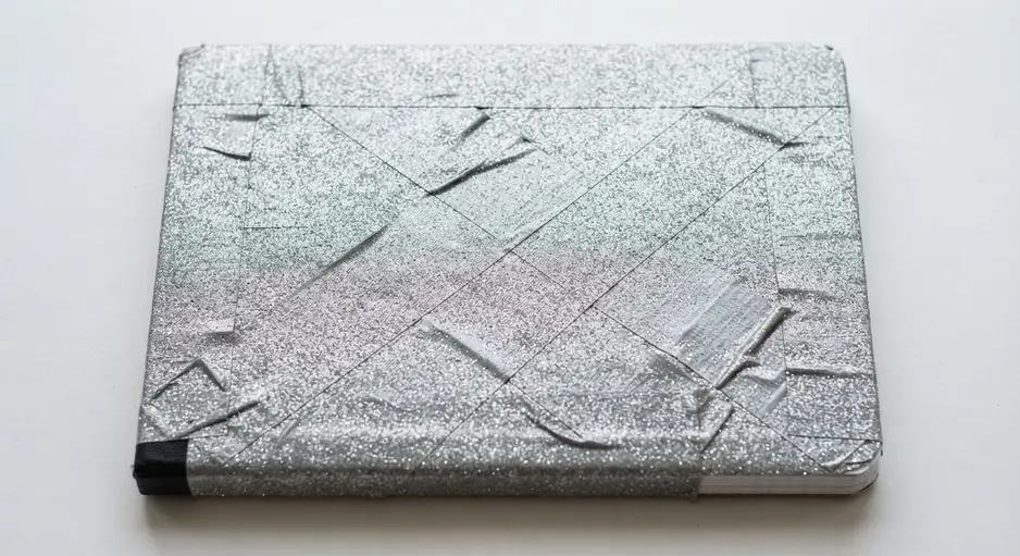 A standard composition notebook with its cover completely recovered in overlapping strips of silver glitter duct tape.