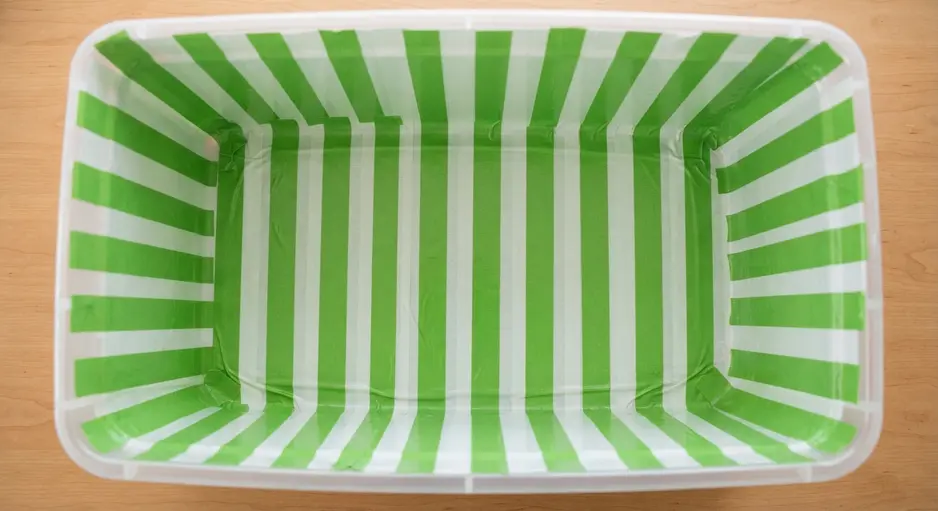 A clear plastic storage bin with its interior lined with vertical stripes of green and white duct tape.