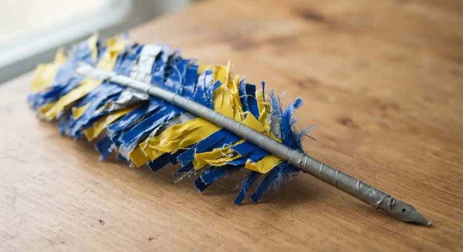 A feather quill shape attached to a pen, created by cutting and layering fringed strips of bright blue and yellow duct tape.