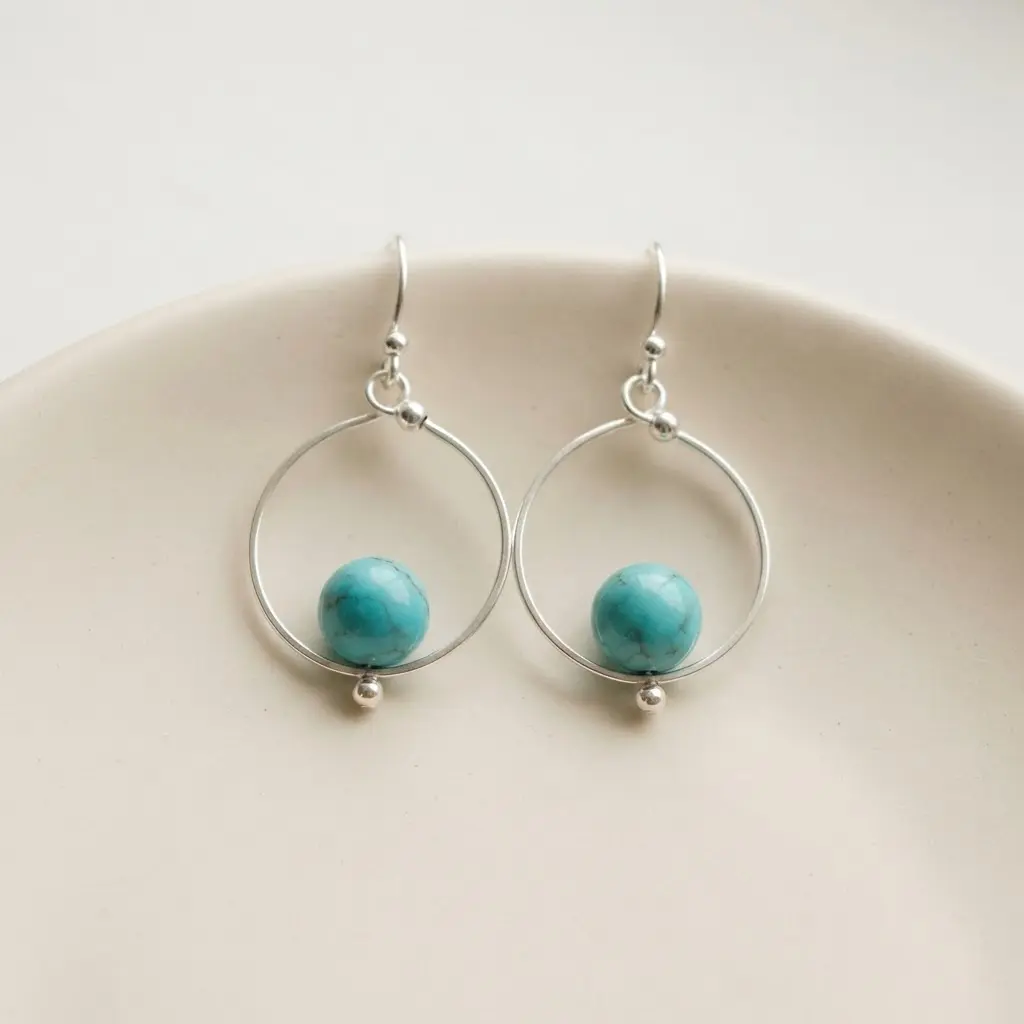 A pair of small silver hoop earrings with a single round turquoise bead hanging from the bottom.