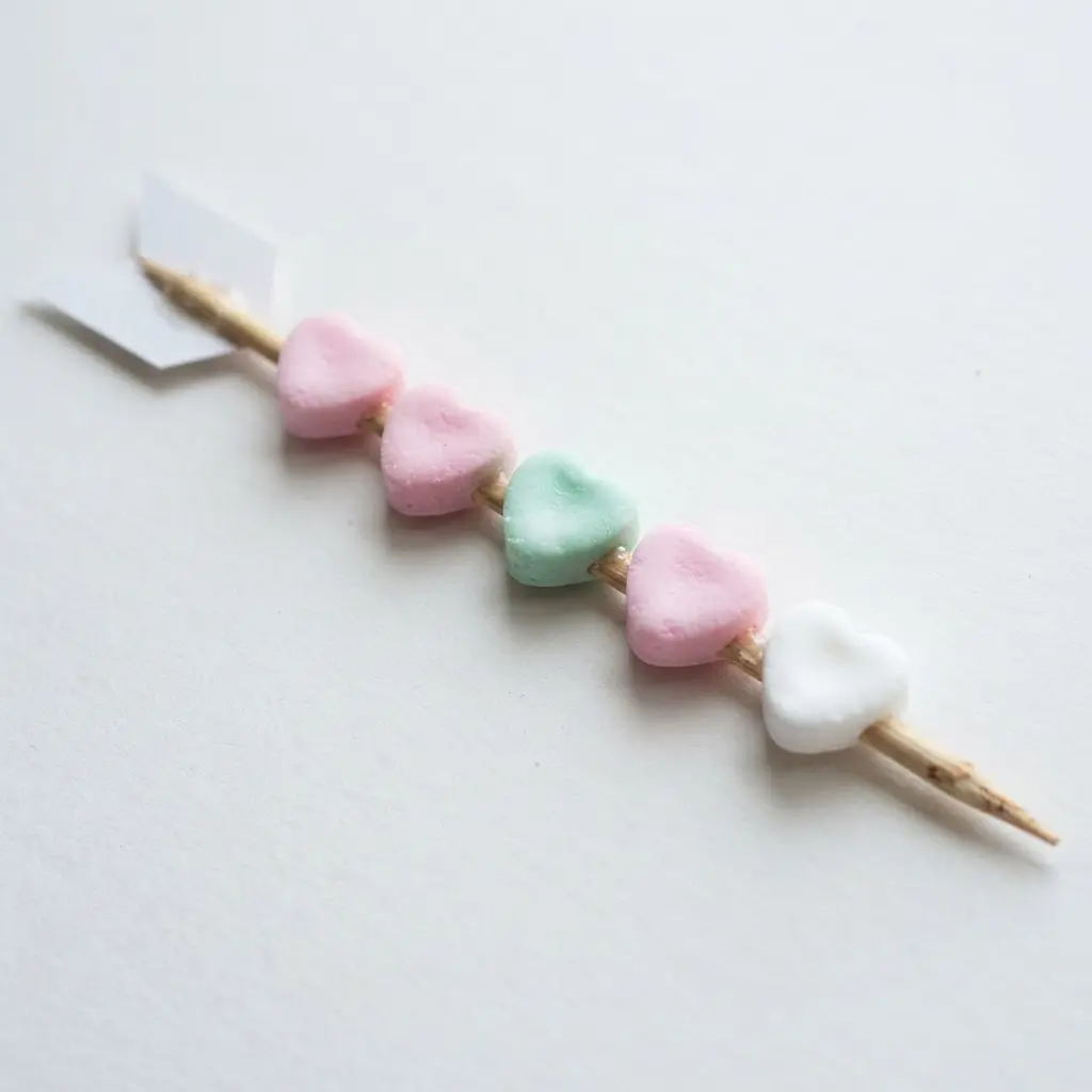 A pink and white arrow made from a wooden skewer with pastel conversation hearts glued along the shaft and at the tip.