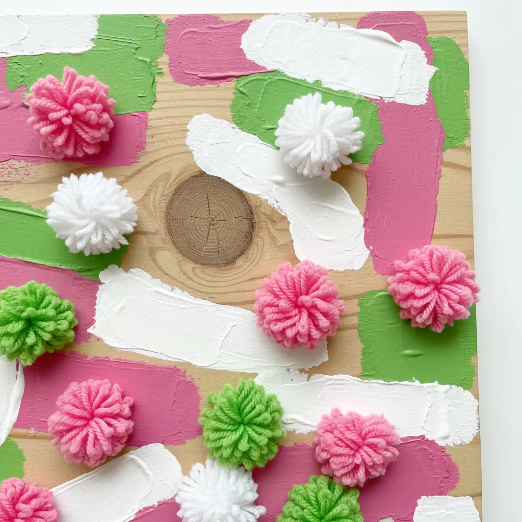 A small wooden sign reading 'Create' in white, with a child's hand gluing on fluffy purple and green pom poms along the border.