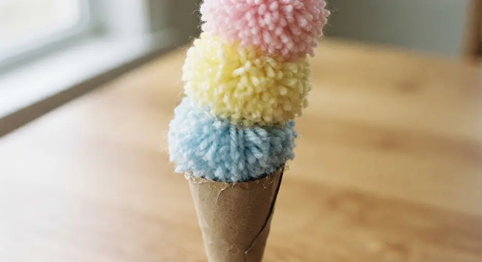 A tall cardboard cone topped with a stack of pink, yellow, and blue fluffy yarn pom poms.