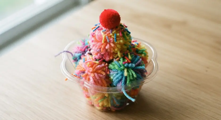 A clear plastic dish filled with rainbow-colored yarn pom pom scoops, yarn sprinkles, and a red pom pom cherry.
