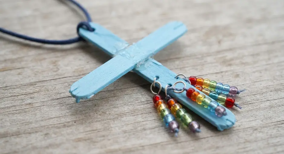 A light blue painted cross made from two sticks with multi-colored bead fringe hanging from the bottom.