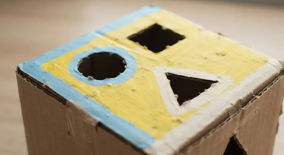 A child's hand dropping a yellow wooden triangle block through a matching hole in a painted cardboard box.
