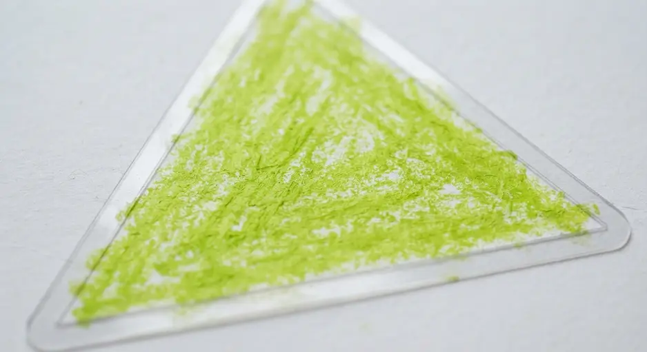 A child using a green crayon to trace the inside of a plastic heart stencil held steady on white paper.