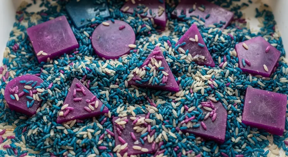 A child's hands sifting through blue-dyed rice in a plastic bin to uncover a large purple plastic hexagon.