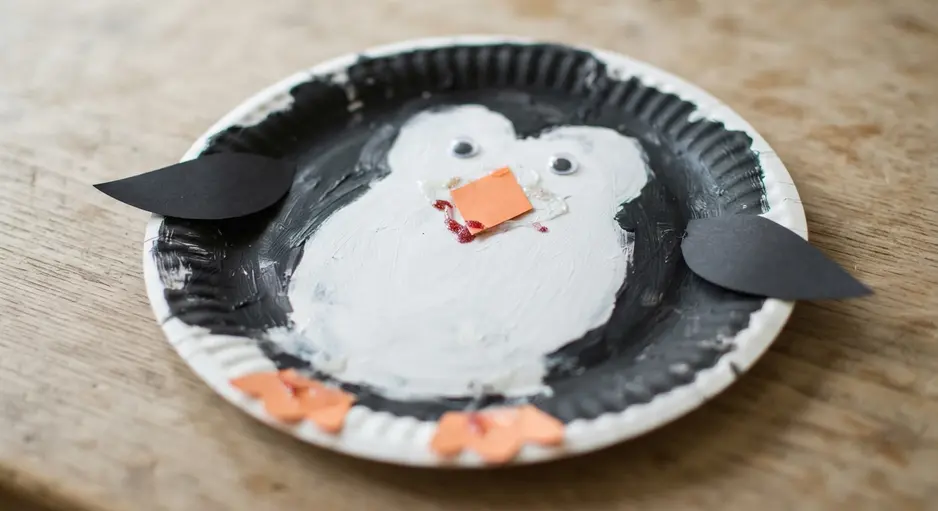 A black and white penguin craft with a paper plate body, orange beak, and googly eyes.